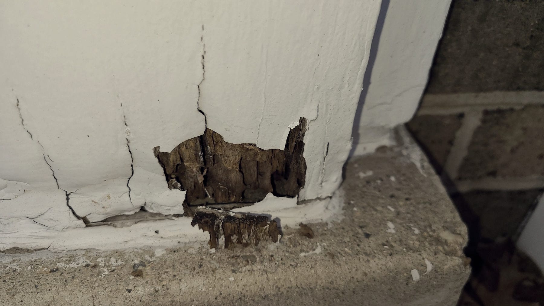 Damage where a wall meets the floor beside a baseboard, with a broken section of the wall surface and debris on the floor.
